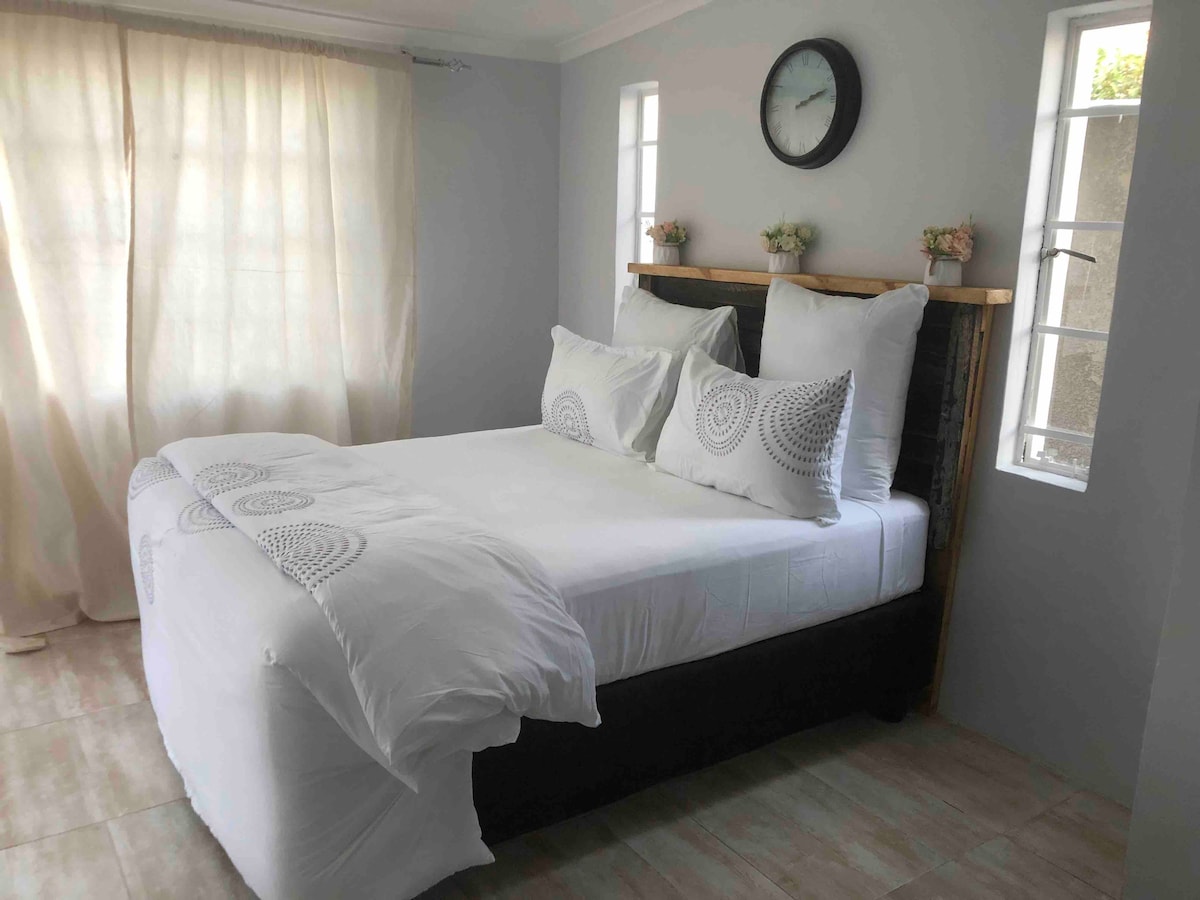 A bright sleeping area features a spacious bed adorned with white linens and decorative pillows. Light streams in from multiple windows, complemented by soft curtains. A clock is placed on the wall above the bed, adding a touch of charm to the minimalist design.