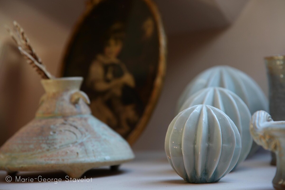 A collection of decorative porcelain items is displayed on a shelf. The pieces include a bulbous vase with a textured finish, two spherical ornaments with ridged surfaces, and a small curved bowl. A framed portrait partially visible in the background adds a touch of charm.