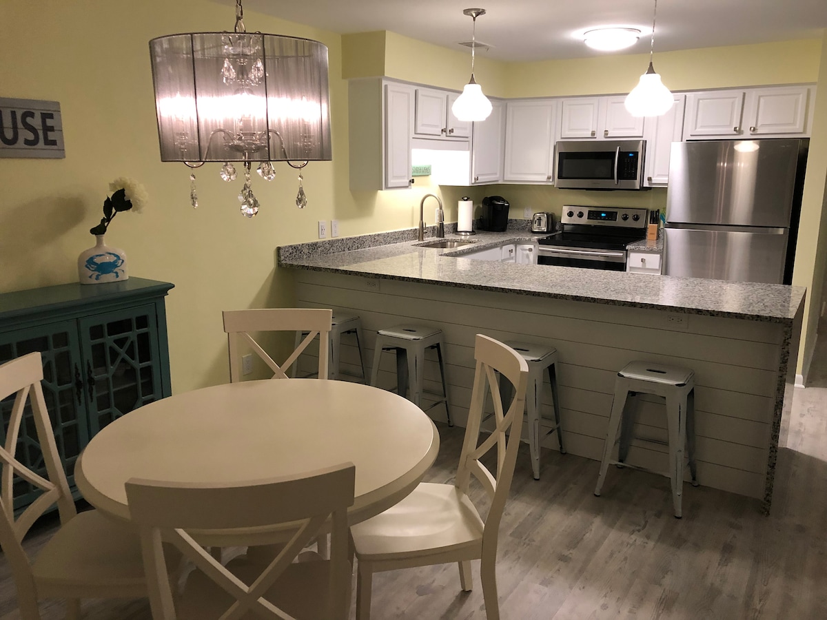 A modern kitchen and dining area is presented with light green walls and granite countertops. White cabinetry and stainless steel appliances complement the space. A round dining table surrounded by four chairs is positioned next to a decorative cabinet. Two barstools are placed at the kitchen counter.