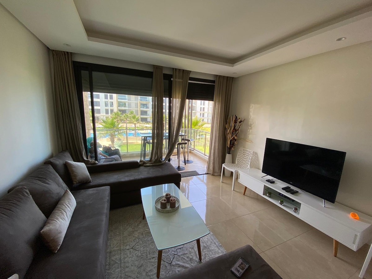 The living room features a spacious sofa arranged for comfort and relaxation, accompanied by a coffee table. Large windows with sheer curtains offer views of the patio and pool area, allowing natural light to illuminate the space. A television is mounted on a sleek white unit.