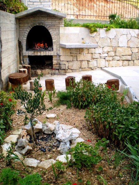 An outdoor space features a stone barbecue area with a brick oven. Surrounding greenery includes shrubs, small plants, and decorative stones arranged in a circular pattern. Wooden seating is provided around a fire pit, enhancing the inviting atmosphere of the garden.