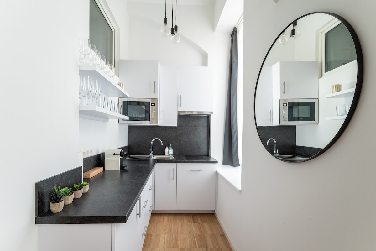 A modern kitchen with white cabinetry is complemented by a dark countertop. Two microwaves are positioned above a sleek sink, while a circular mirror reflects the natural light from the window. Small potted plants are neatly arranged along the counter.