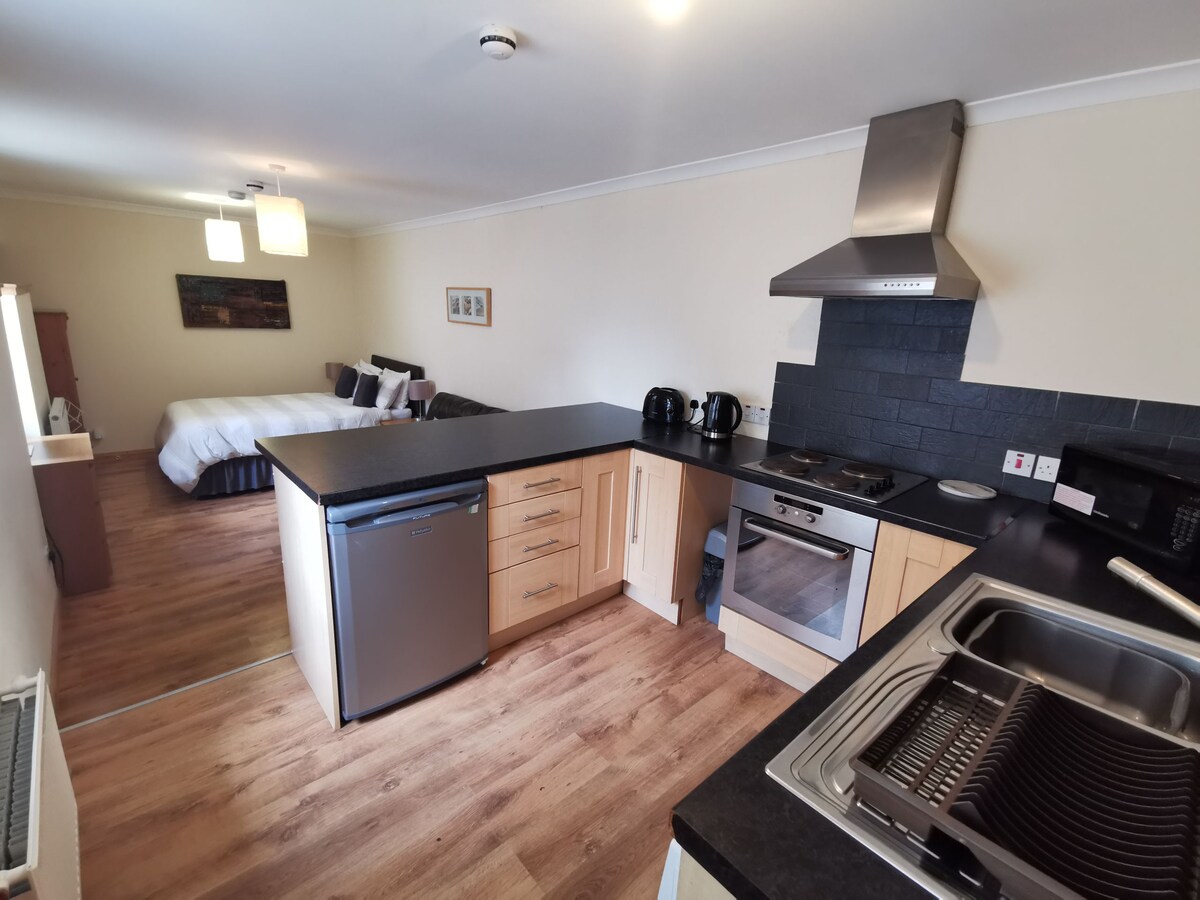 The fully equipped kitchen features modern appliances, including a stove, oven, and refrigerator. A countertop separates the kitchen from the sleeping area, where a double bed is visible in the background. Natural light fills the space, highlighting the warm wooden flooring.
