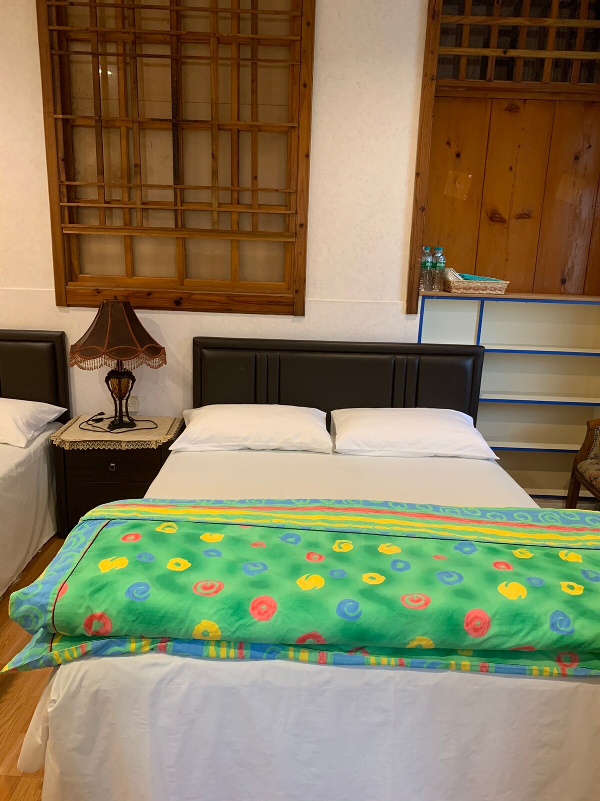 A comfortable sleeping area is presented, featuring a spacious bed with white linens and a colorful blanket adorned with floral patterns. A bedside lamp adds illumination next to a small table, while shelving is visible in the background alongside wooden paneling.