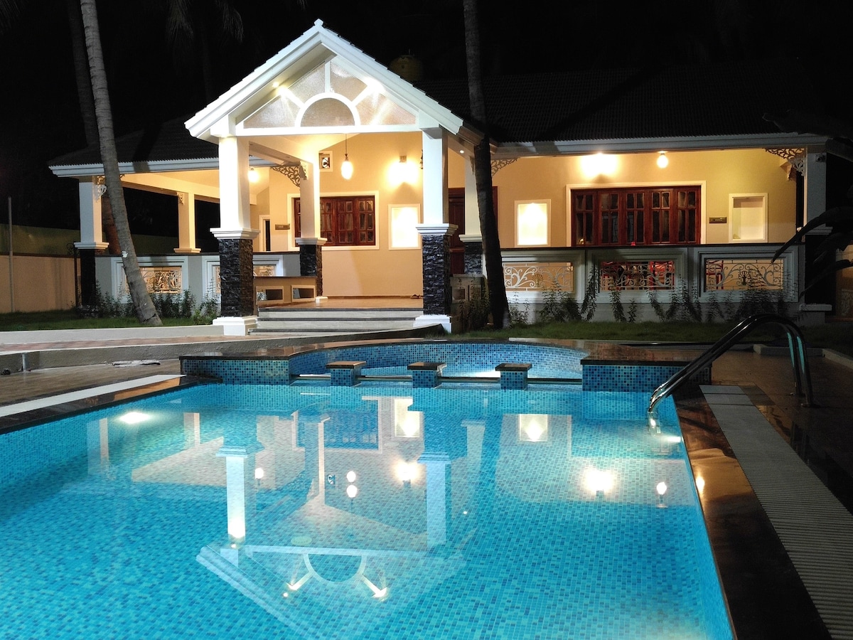 A spacious farmhouse is showcased under soft evening lighting, with a private pool reflecting the structure. The pool area features a tiled edge and steps leading into the water. The house, with its elegant architectural details, is framed by palm trees.