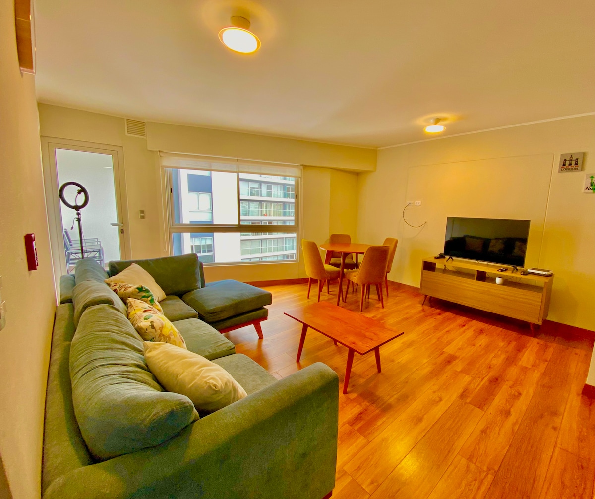 A spacious living area features a large green sectional sofa and a wooden coffee table. A round dining table with upholstered chairs is positioned nearby. Natural light enters through large windows, illuminating the warm wood flooring. A wall-mounted TV is visible in the corner.