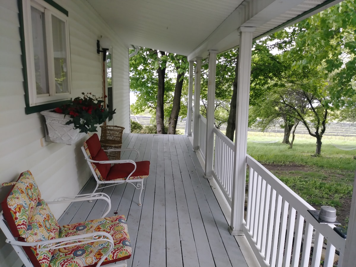 A covered porch is lined with white railings and features two chairs with colorful cushions. Potted plants are arranged on one side, while trees offer shade and a view of the surrounding landscape. A serene atmosphere is created by the peaceful outdoor setting.
