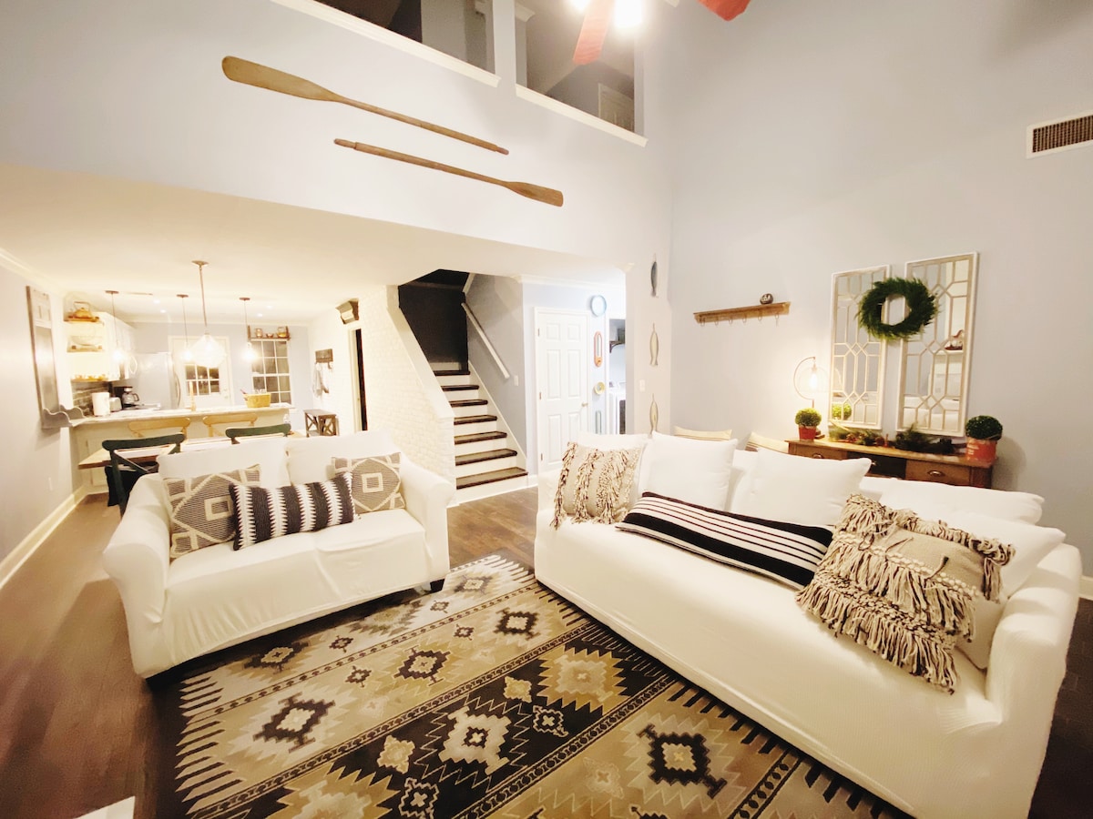 A spacious living area features two white sofas adorned with patterned pillows, arranged around a textured area rug. Natural light illuminates the room, highlighting the warm wood floors and the open concept design that connects to the dining area and kitchen.