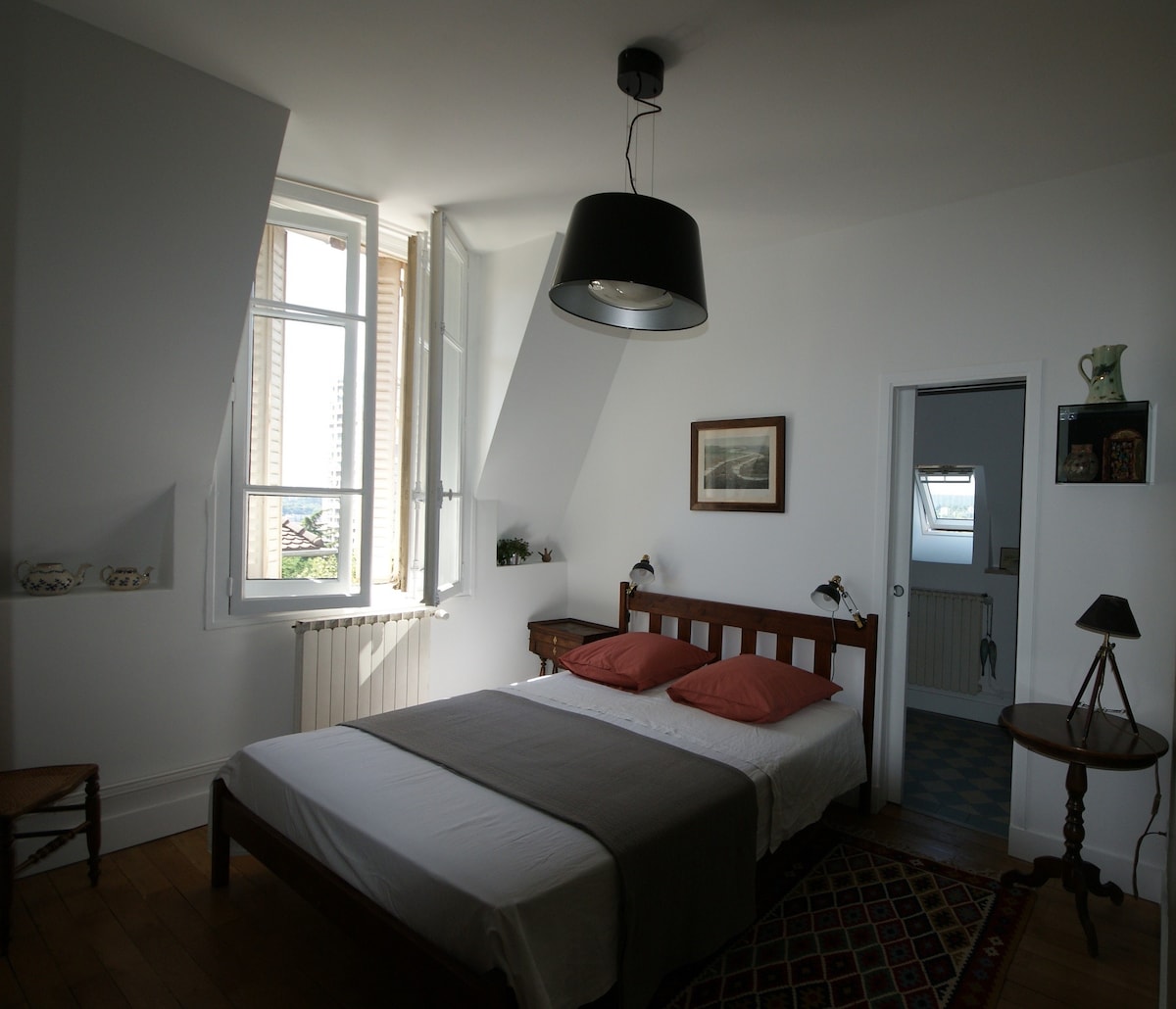 The room features a spacious bed with orange accent pillows. Large windows allow natural light to fill the area, while a warm wooden floor complements the decor. A simple desk lamp and artwork enhance the minimalist style, providing a calm atmosphere.