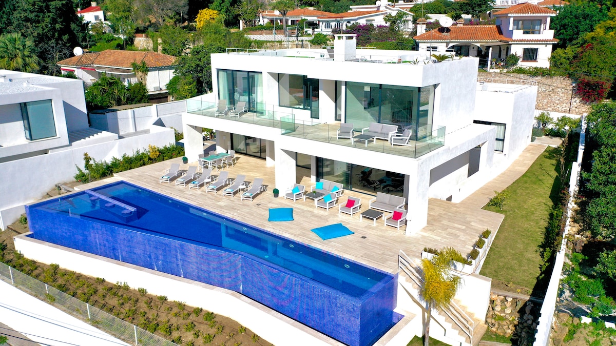 An aerial view of Villa Infinity illustrates a contemporary design with expansive glass windows. A large infinity pool stretches across the terrace, surrounded by sun loungers in various colors. Lush greenery and landscaped gardens complement the villa's structure.