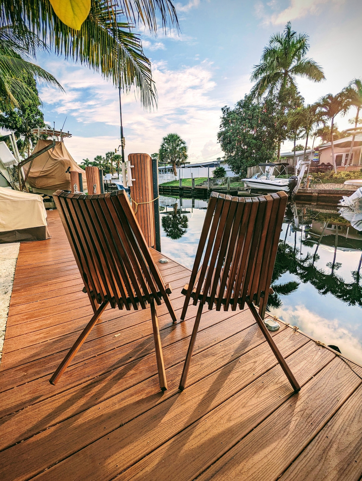 Two wooden chairs sit on a wooden deck by the water's edge, reflecting calmness in the surrounding water. Palm trees provide a tropical backdrop under the bright sky, with scattered clouds adding texture to the serene atmosphere.