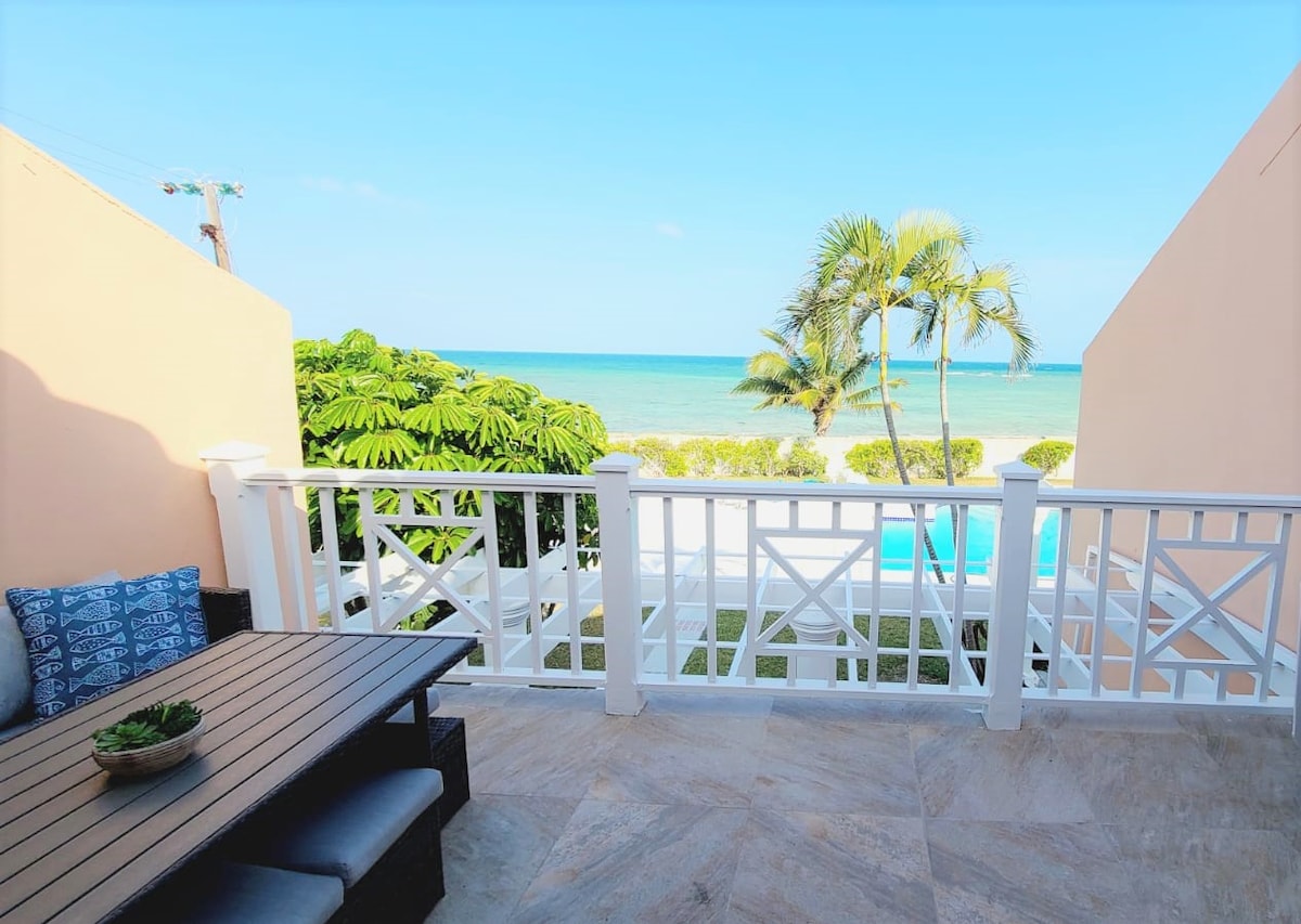 An outdoor balcony setting features a wooden dining table surrounded by comfortable seating. Lush greenery and palm trees are visible below, with a scenic view of the beach and ocean in the distance under a clear blue sky.