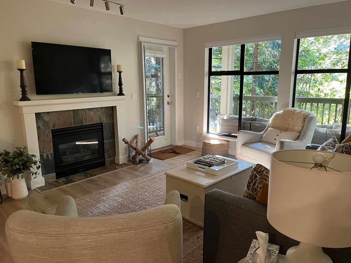 A welcoming living area features a light-colored sofa and armchairs, complemented by a decorative coffee table. A flat-screen TV is mounted above a stone fireplace, while large windows provide views of the outdoors, allowing natural light to fill the space.