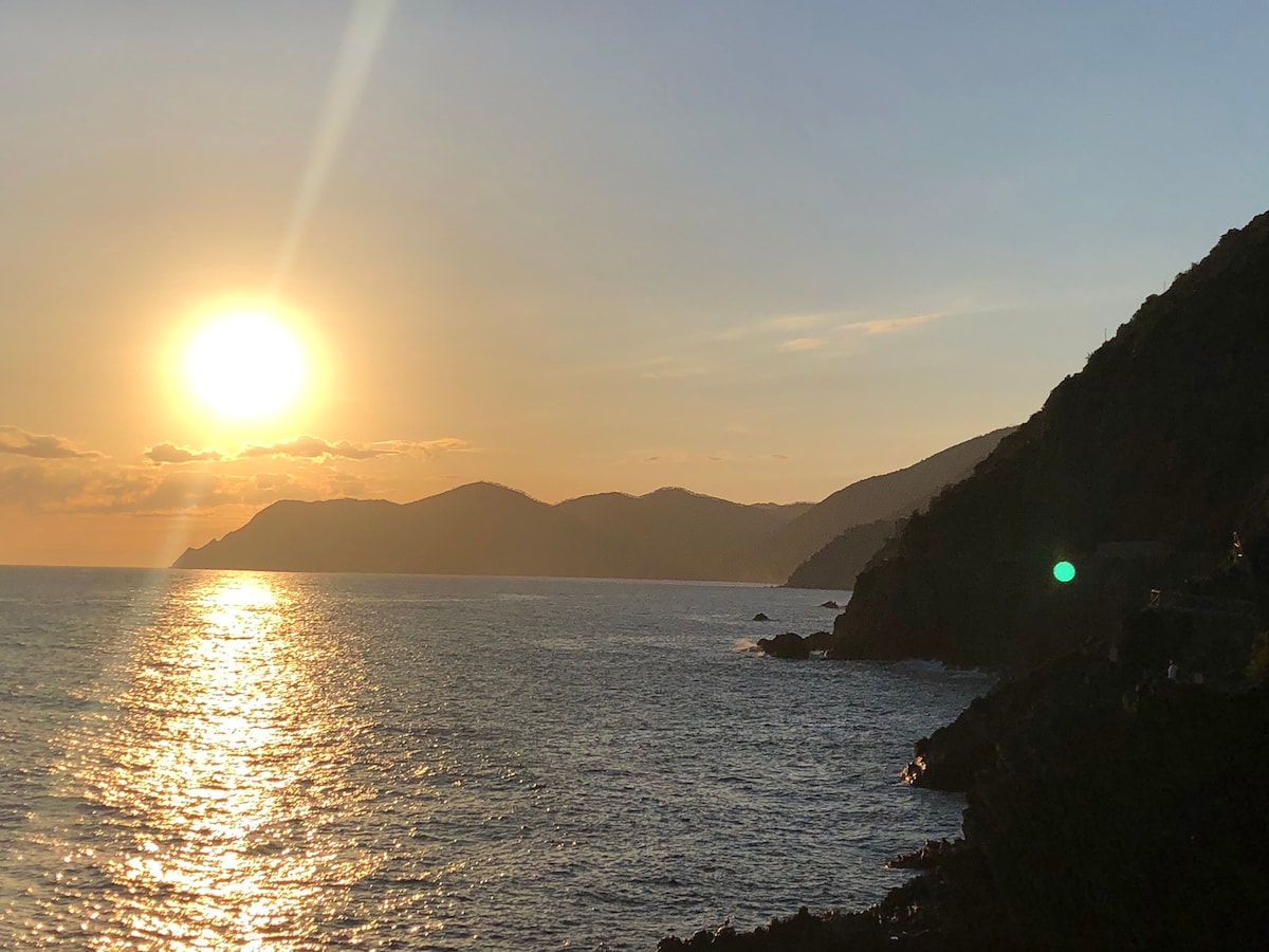 A sunset is observed over the shimmering sea, with golden rays reflecting on the water's surface. The silhouette of mountains provides a backdrop, adding depth to the tranquil scene. The sky transitions from warm oranges to soft blues, enhancing the serene atmosphere.