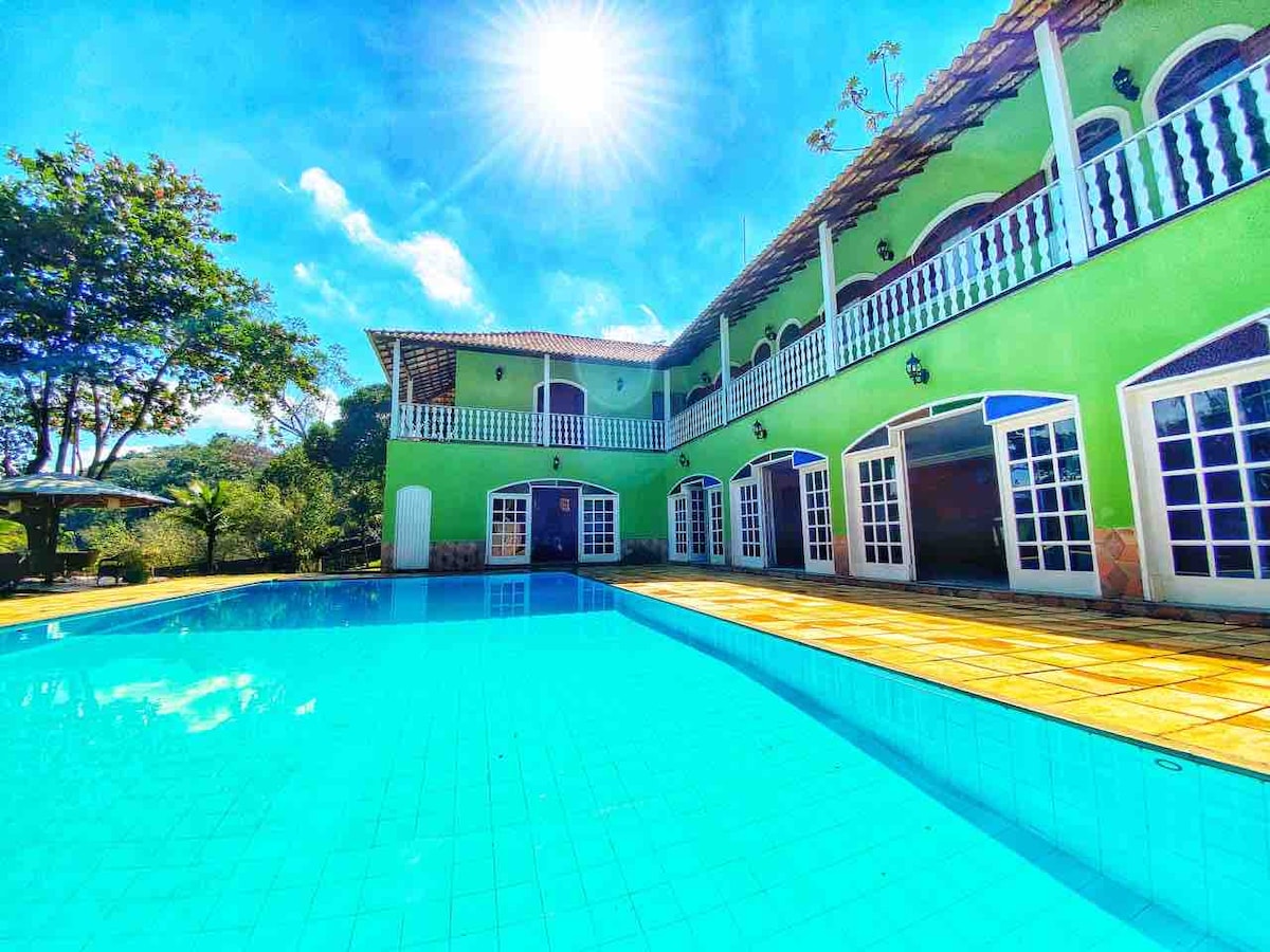 A vibrant green building is viewed beside a spacious pool, reflecting sunlight. Large glass doors open onto the patio area, enhancing the connection to the outdoors. Clear blue skies and surrounding trees create a serene backdrop.
