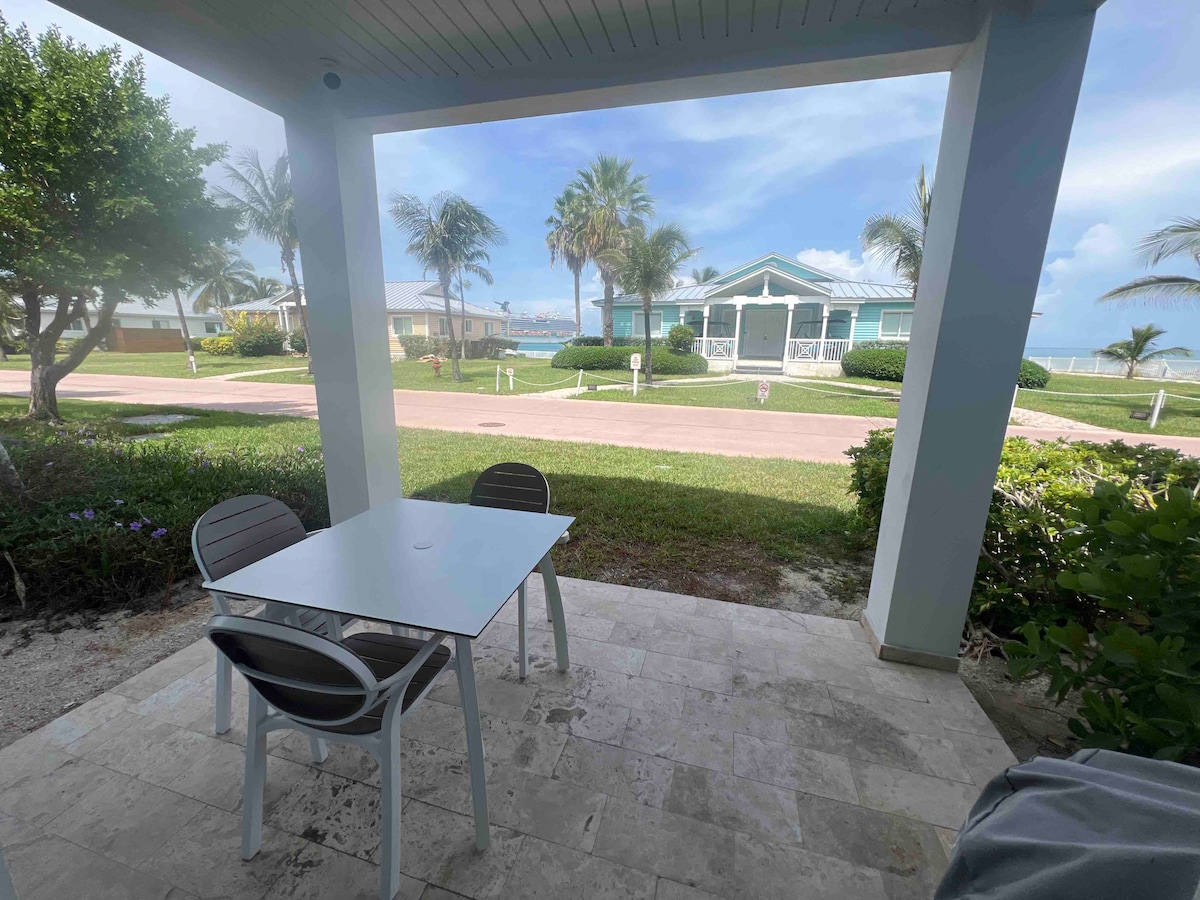 A patio area features a small outdoor table with four chairs, surrounded by greenery. The view extends to a quiet street lined with palm trees and homes, with glimpses of the ocean in the distance under a clear blue sky.
