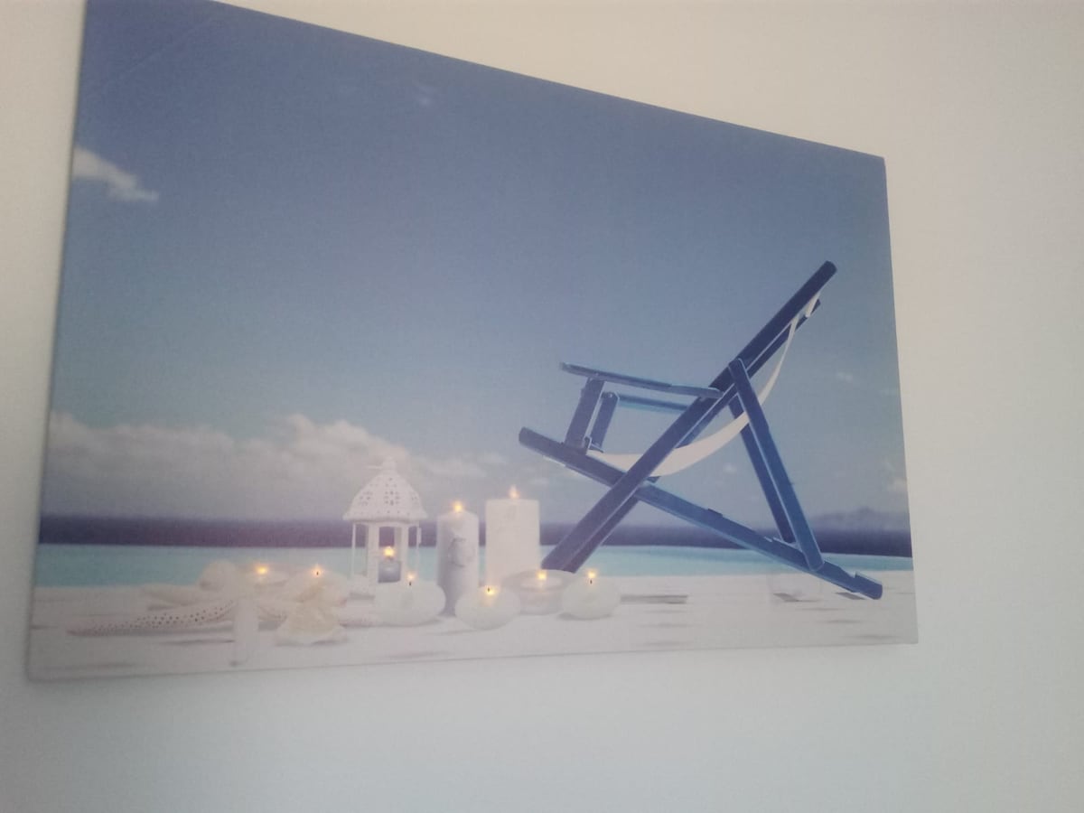 A wall-mounted artwork depicts a serene coastal scene, featuring a blue chair beside white stones and lanterns. Soft white candles are arranged prominently in the foreground, evoking a calm and relaxed atmosphere against a backdrop of a clear blue sky and gentle waves.