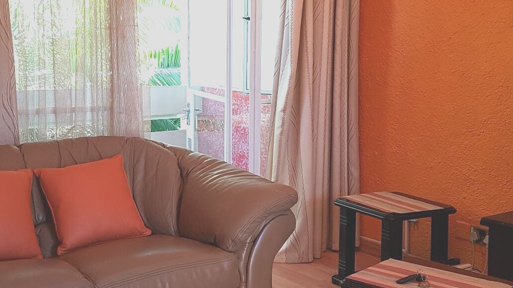 A comfortable lounge area features luxurious leather couches with bright orange cushions, inviting seating arrangements, and small coffee tables. Light filters through sheer curtains, creating a warm ambiance. A balcony is visible through the partially open sliding door, enhancing the space's openness.