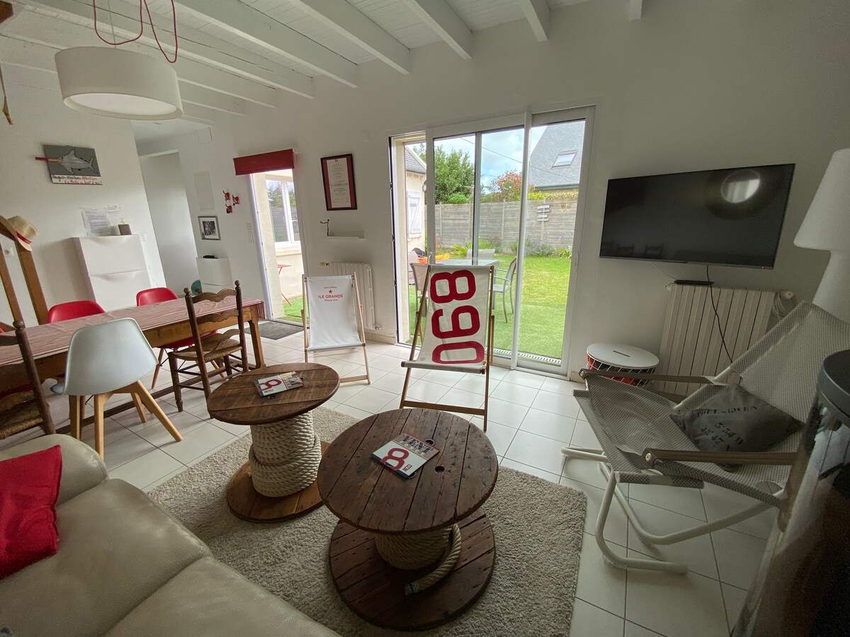 A bright living area features two wooden coffee tables and neutral-toned seating. Glass doors provide access to the garden, while a wall-mounted television is visible. A cozy rug anchors the space, and decorative accents in red complement the light color palette.