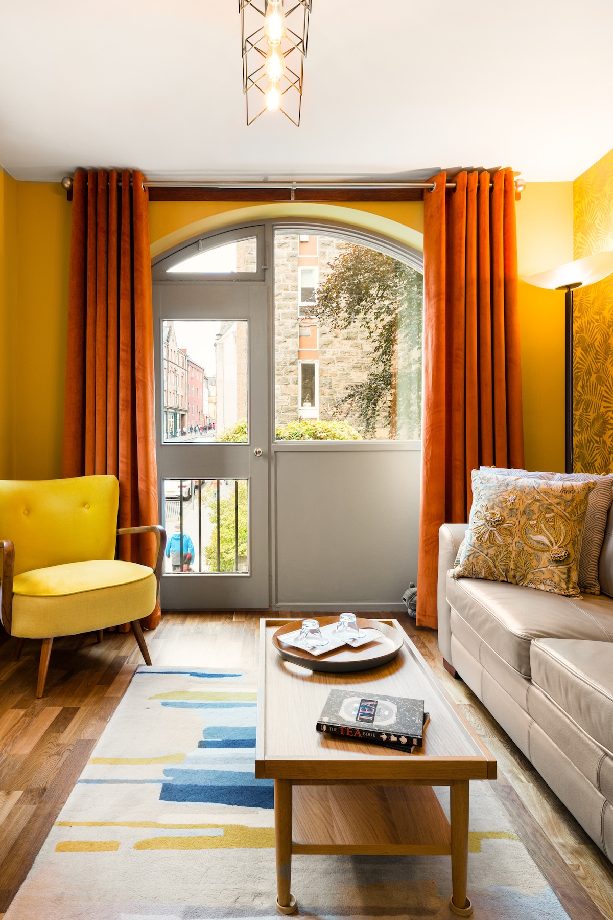 A well-lit lounge features a large arched window draped with rich orange curtains, overlooking the street. A cozy yellow armchair complements a neutral sofa adorned with patterned cushions. A wooden coffee table sits in front, displaying a book and glassware, on a colorful area rug.