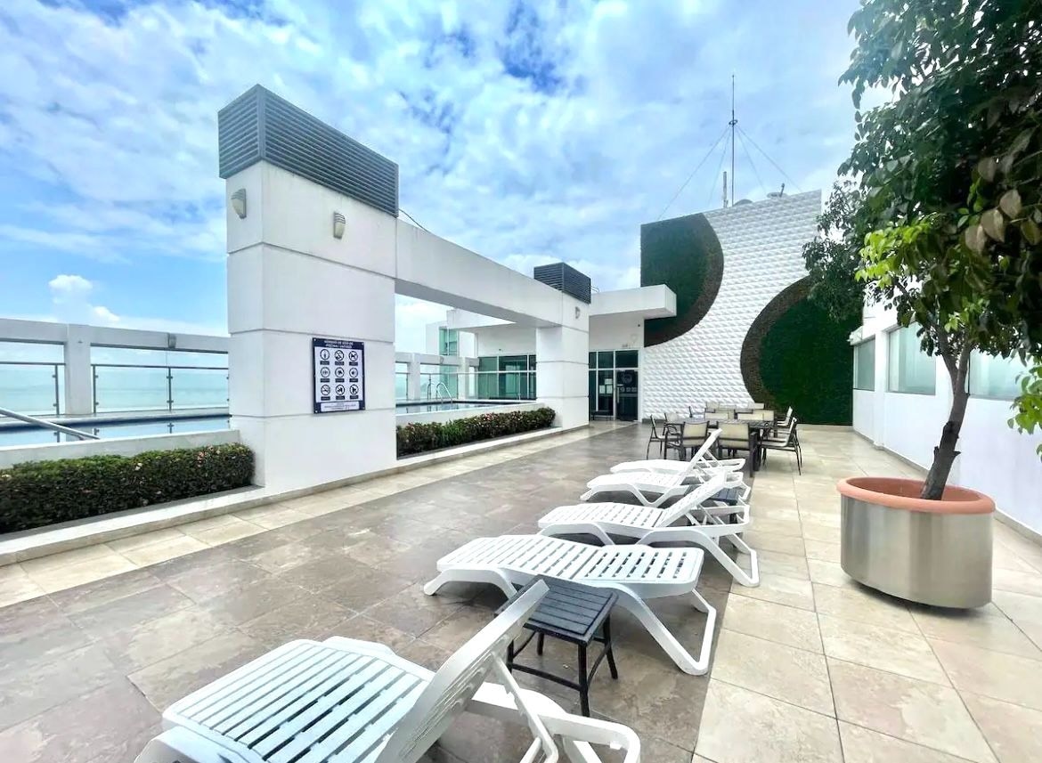 A spacious rooftop area features several lounge chairs arranged along the edge, with a large dining table placed nearby. Lush greenery is visible alongside the building, and the sky above is partly cloudy, allowing natural light to illuminate the space.