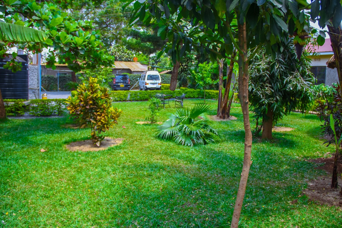 A landscaped garden features a mix of lush green grass and various plants. Several trees provide shade, while a couple of seating areas are visible among the vegetation. The surroundings are well-maintained, creating a peaceful outdoor space.
