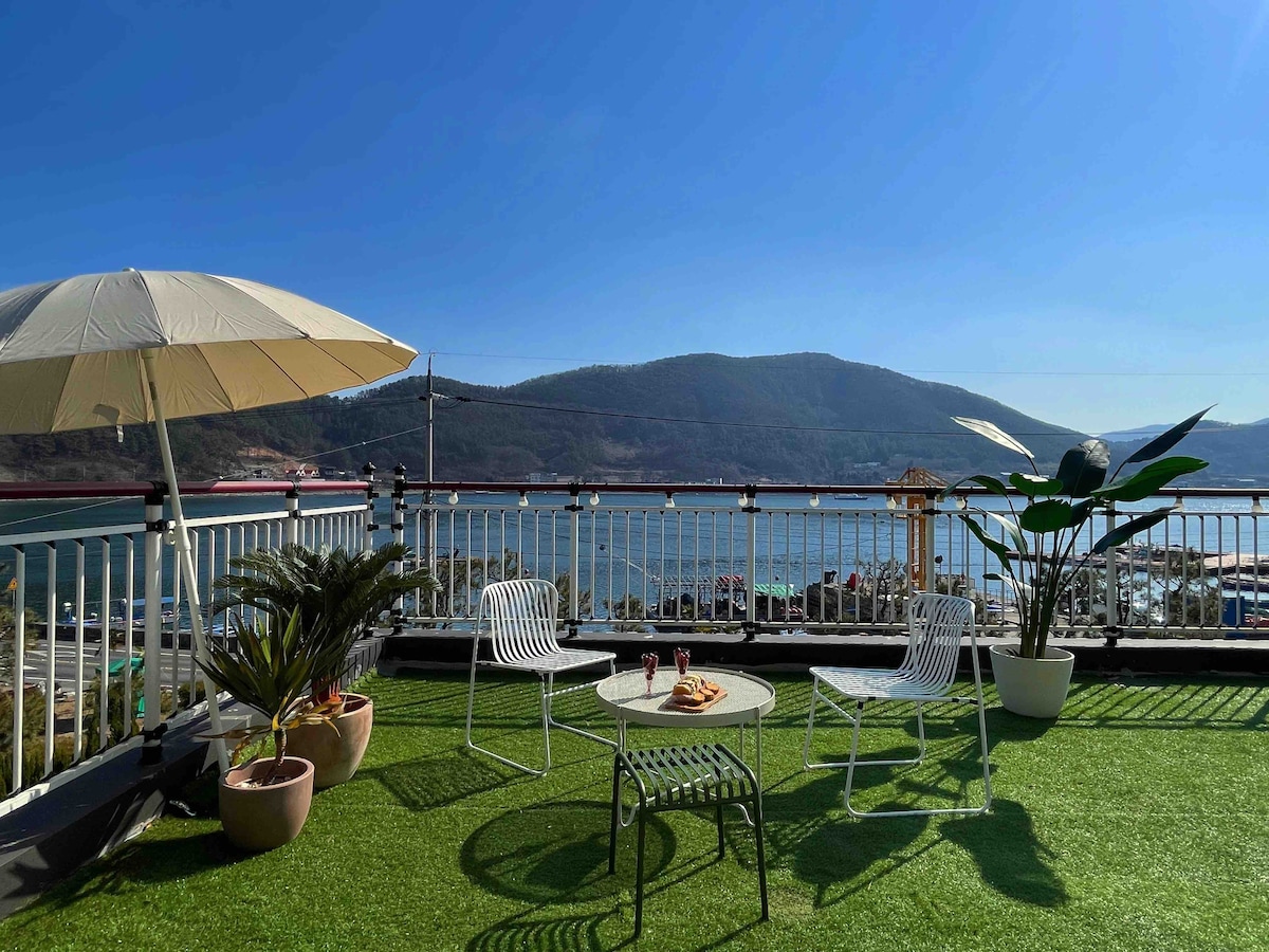 A spacious terrace is shown with views of the ocean and mountains. Two white chairs and a small table are seen alongside potted plants. An umbrella provides shade, and bright sunlight illuminates the scene, emphasizing the inviting outdoor space.