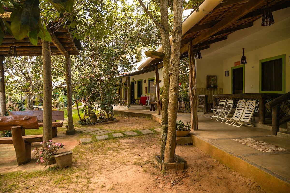 A serene outdoor space is surrounded by trees, with paths leading between simple structures. Seating areas are located on porches, offering views of the lush greenery. A variety of plants are present, enhancing the peaceful garden atmosphere.
