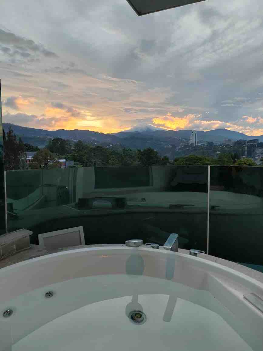 A spacious hot tub is positioned next to a glass railing, offering uninterrupted views of the distant mountains. The sky is filled with soft hues of orange and pink as the sun sets, creating a serene backdrop for relaxation.