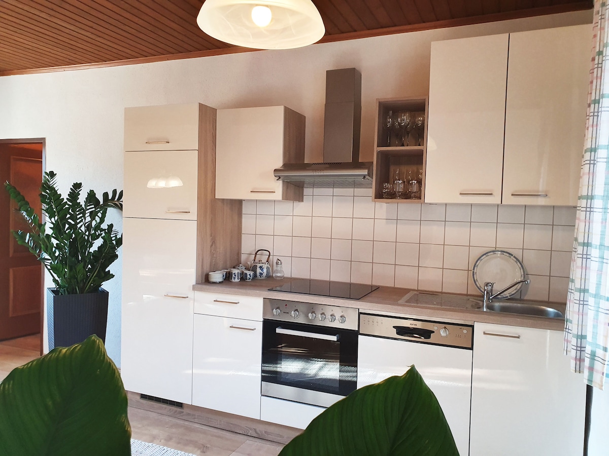 A modern kitchen features sleek white cabinetry paired with wooden accents. An oven, stovetop, and sink are integrated along the counter. A large potted plant adds greenery, while natural light filters through a nearby window, creating a welcoming environment for meal preparation.