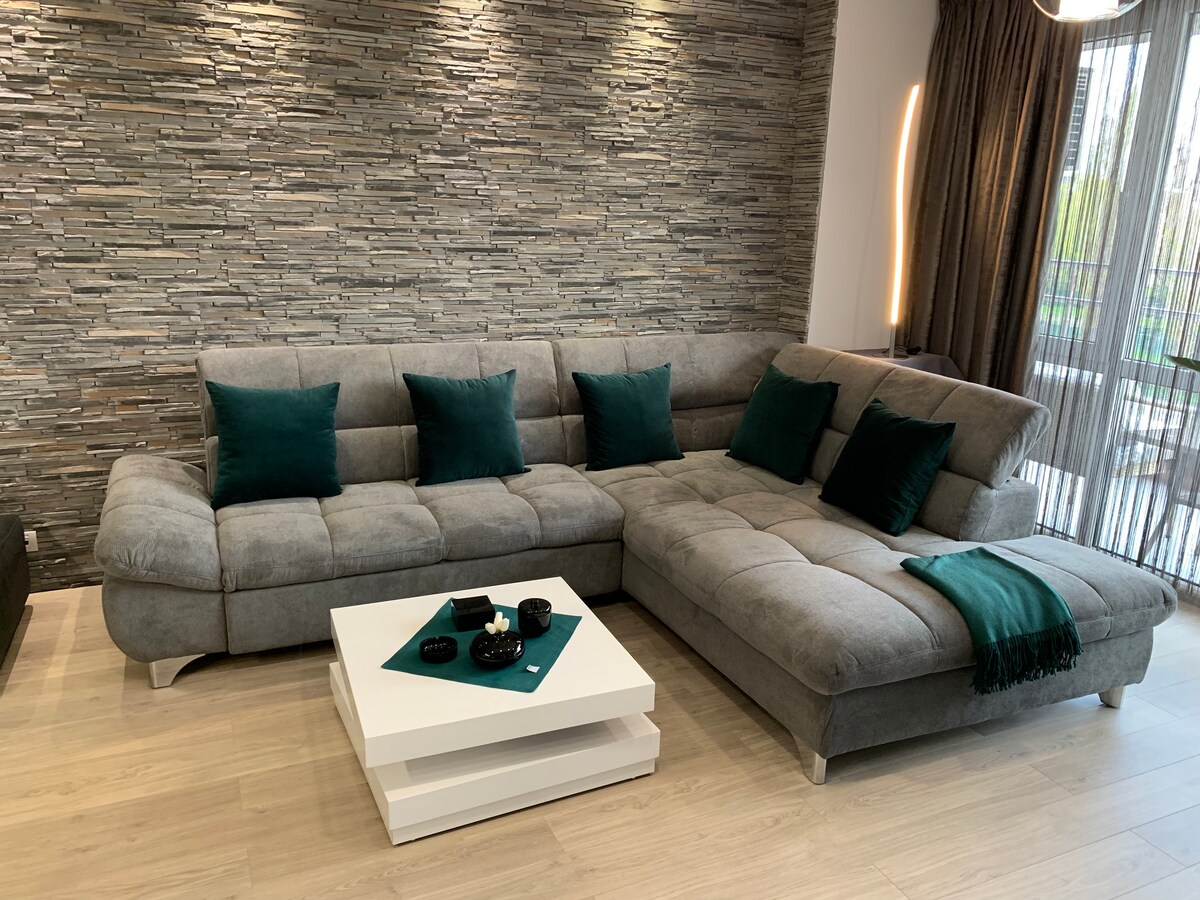A spacious gray sectional sofa is arranged thoughtfully against a textured stone wall, complemented by dark green decorative pillows. A modern white coffee table sits in front, adorned with candles and a small decorative tray, while natural light filters through sheer curtains.