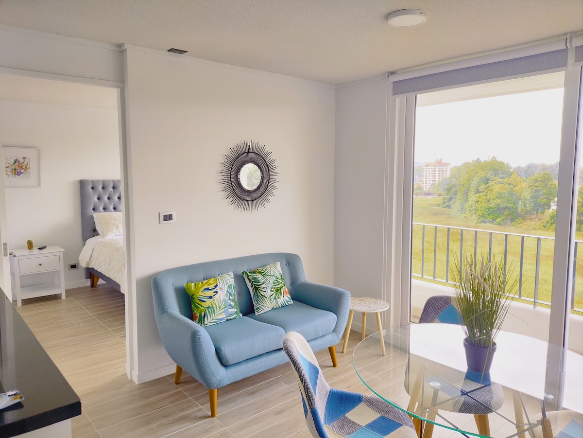A light-filled living area features a modern blue sofa with tropical-patterned cushions, paired with a circular glass dining table. A mirror is mounted on the wall, reflecting the fresh colors. Large windows offer views of the surrounding greenery, enhancing the space's openness.