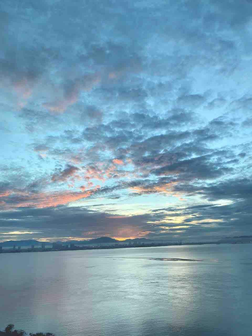 A serene view of the sea is presented, reflecting the colors of a sunset sky filled with clouds. Soft hues of orange and pink blend with deeper blue tones, creating a calming atmosphere over the water's surface, which appears smooth and undisturbed.