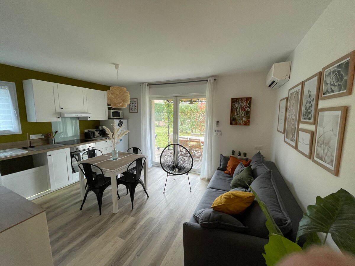 A cozy living space features a comfortable gray sofa accented with bright pillows, a dining area with a white table and black chairs, and a fully equipped kitchen. Large glass doors open to a deck, allowing natural light to fill the room and connecting to the outdoor scenery.