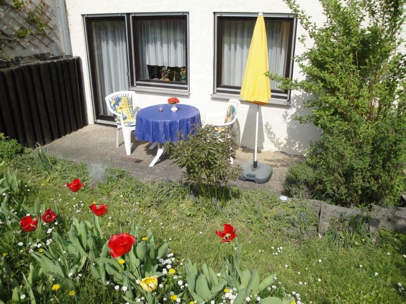 An outdoor terrace features a round blue table with two white chairs, accompanied by a yellow umbrella. Colorful tulips and greenery surround the area, enhancing the garden view. Large windows of the apartment are visible in the background, allowing natural light into the indoor space.