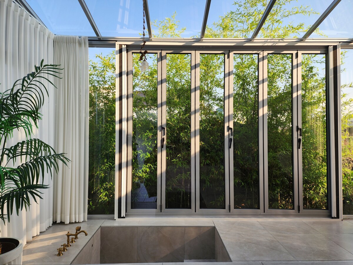 A sunroom features large glass doors that open to a lush green view. Natural light floods the space, highlighting a sleek jacuzzi embedded in a modern concrete structure. Soft white curtains frame the scene, adding a touch of elegance to the tranquil setting.