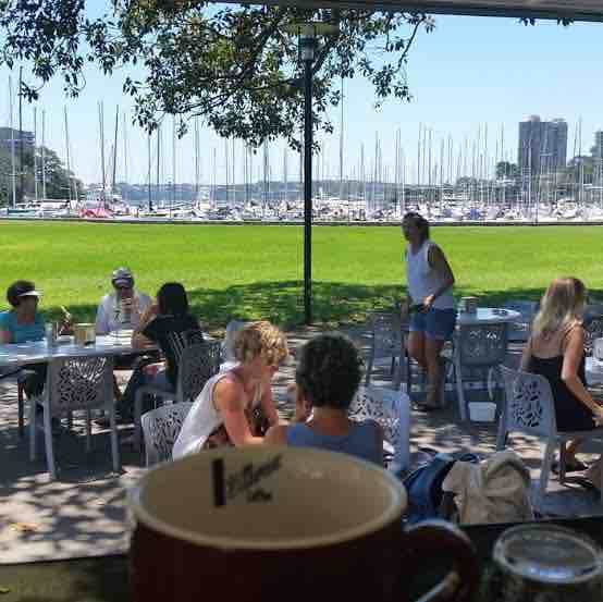 A vibrant outdoor café scene is observed, with patrons seated at modern tables under a shaded structure. A green lawn extends towards a marina, where boats are anchored. The café atmosphere is lively, complemented by the clear blue sky and distant cityscape.