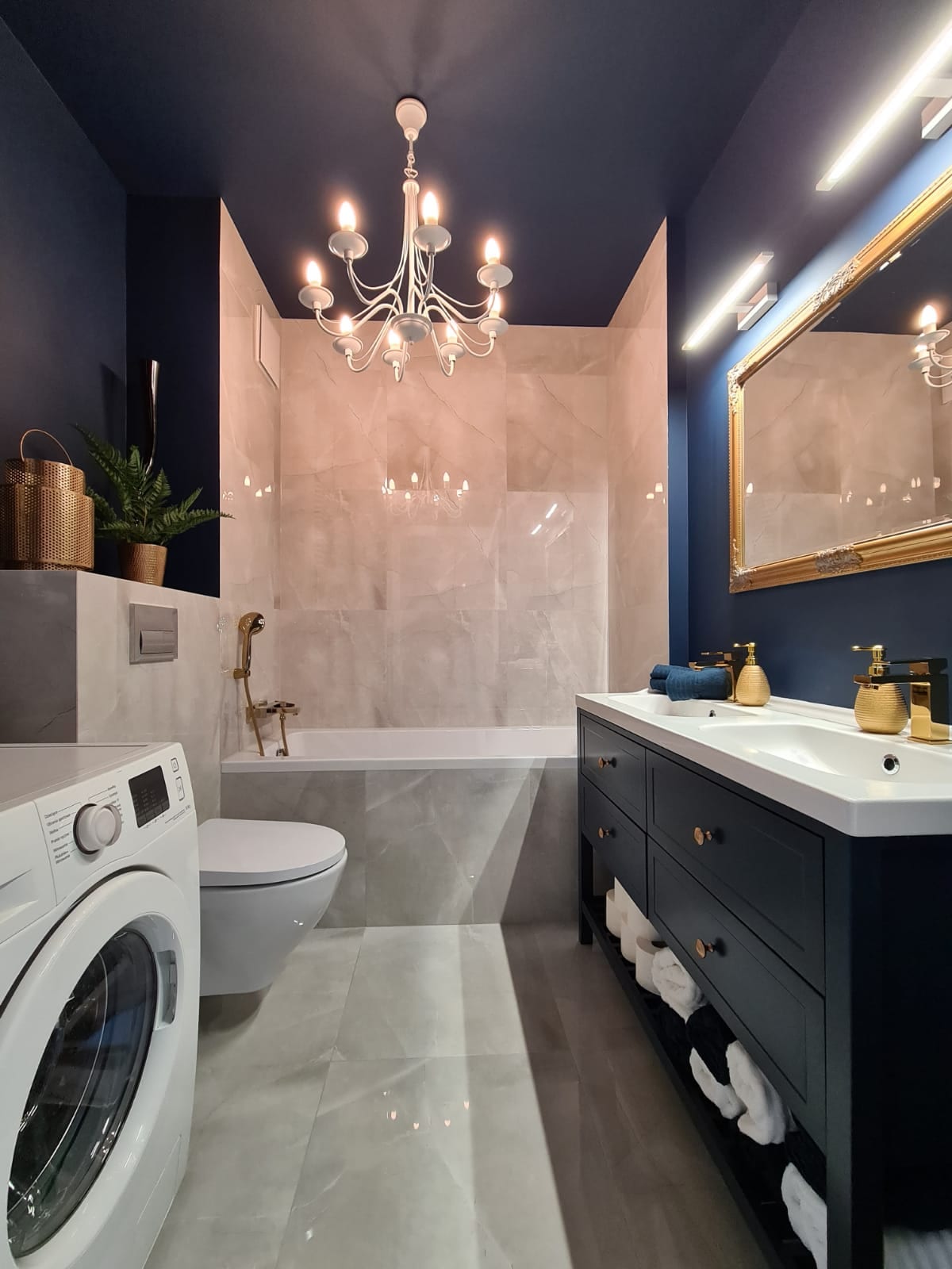 The bathroom features a modern design with a deep bathtub and shower area, both enclosed in sleek tile. A large mirror reflects elegant lighting from a chandelier above. A stylish vanity with a sink and storage sits below, alongside a washing machine, enhancing functionality.