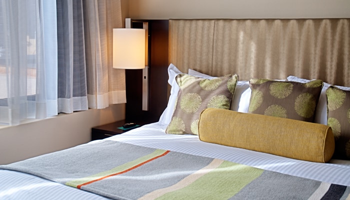 A modern bedroom features a spacious bed adorned with decorative pillows in earthy tones. A soft, colorful blanket adds a touch of warmth. A bedside lamp casts a gentle glow, while sheer curtains allow natural light to filter in, creating a bright yet cozy ambiance.