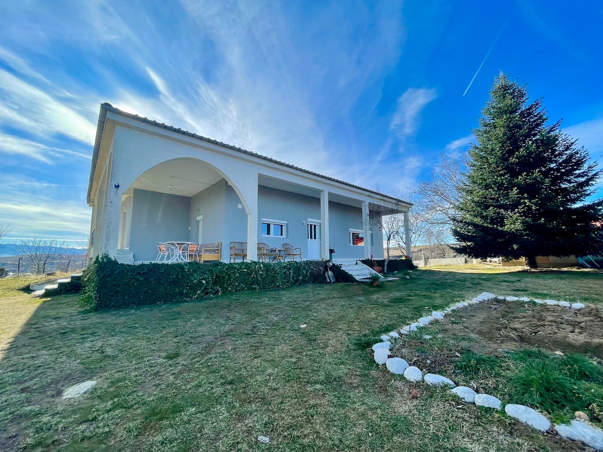 The image depicts a serene farmhouse surrounded by a spacious garden. Features include a large porch with chairs and a table, set against a backdrop of blue sky and distant hills. A tree adds greenery to the landscape, enhancing the peaceful setting.