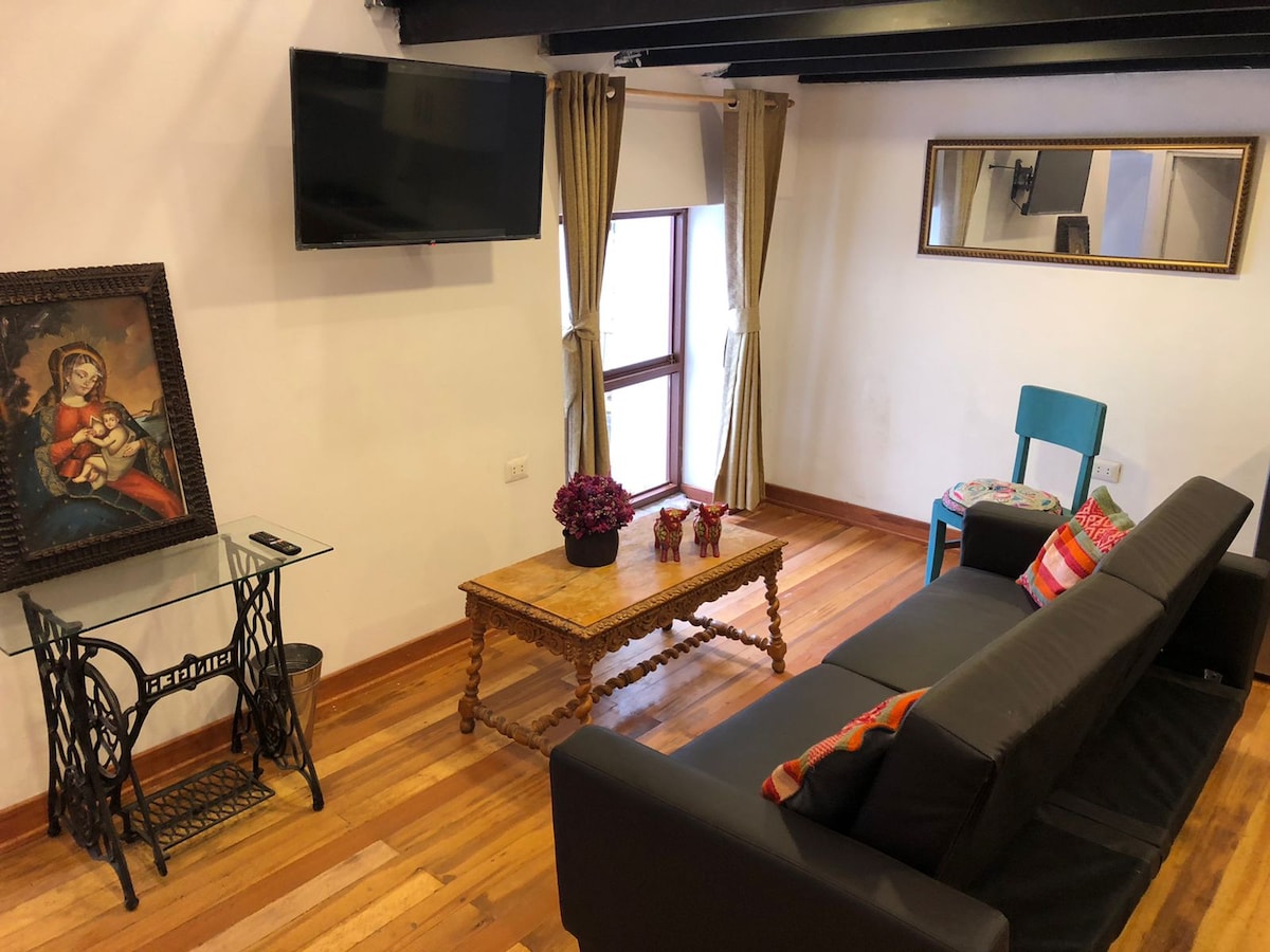 A cozy living area features a dark sofa oriented towards a wall-mounted TV. A wooden coffee table with decorative items is placed nearby. Natural light enters through a window partially covered by curtains. Artwork of a seated figure adds character to the space.
