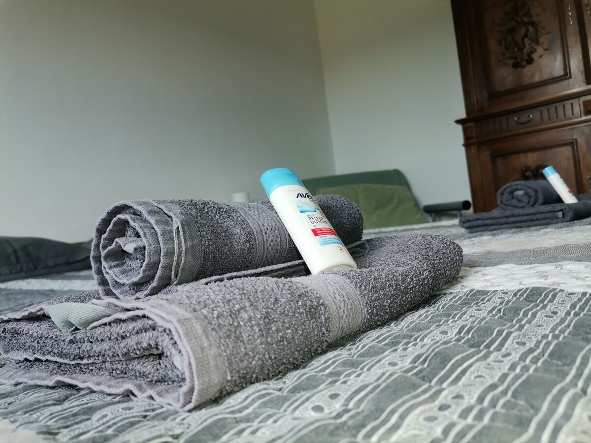 Soft, rolled gray towels are placed neatly on a textured bedding surface. A bottle of lotion is positioned upright nearby. In the background, a green blanket is visible, along with additional rolled towels, all arranged in a calming and organized manner.