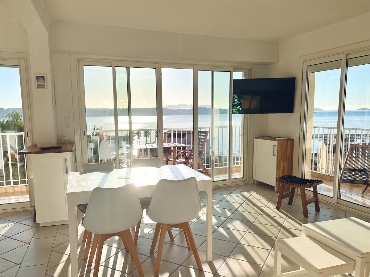 A bright dining area features a white table surrounded by four chairs, with large glass doors providing panoramic views of the bay. Natural light floods the space, illuminating the light-colored tiles and a wooden cabinet. A flat-screen television is mounted on the wall.
