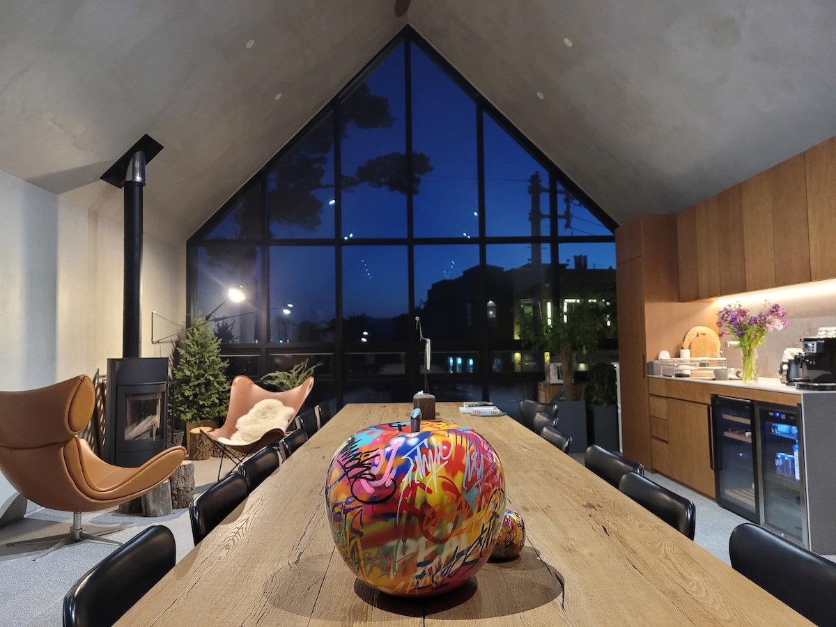 An open dining area showcases a large wooden table at the center, adorned with a colorful decorative piece. A modern kitchen is visible in the background, complemented by a cozy seating arrangement and a large window that frames the evening skyline.