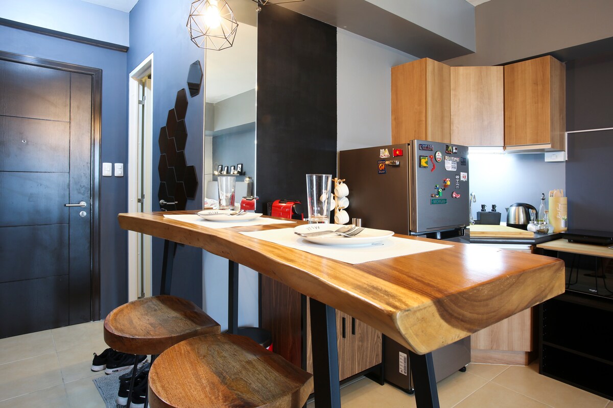 A wooden bar table with two stools is positioned against the wall, complemented by a stylish light fixture overhead. The modern kitchen area features a variety of appliances, including a refrigerator adorned with colorful magnets, and light streaming in from the nearby space.
