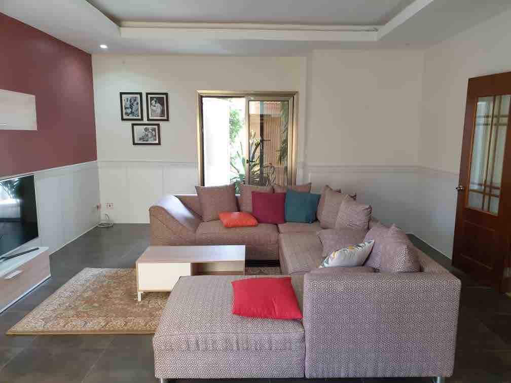 A spacious living area is furnished with a large sectional sofa adorned with colorful cushions. A central coffee table rests on a decorative rug. Natural light enters through a large window, complemented by framed photographs on the wall and a view of indoor greenery.