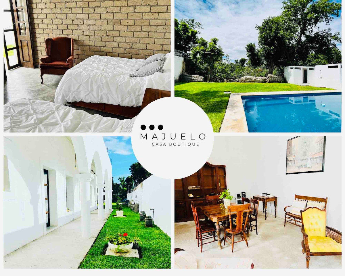 A serene outdoor space features a well-maintained lawn alongside a refreshing pool, surrounded by lush greenery. The interior area displays simple furnishing, including a dining table and chairs set against a backdrop of neutral walls. A cozy bedroom showcases a light quilted bed covering.