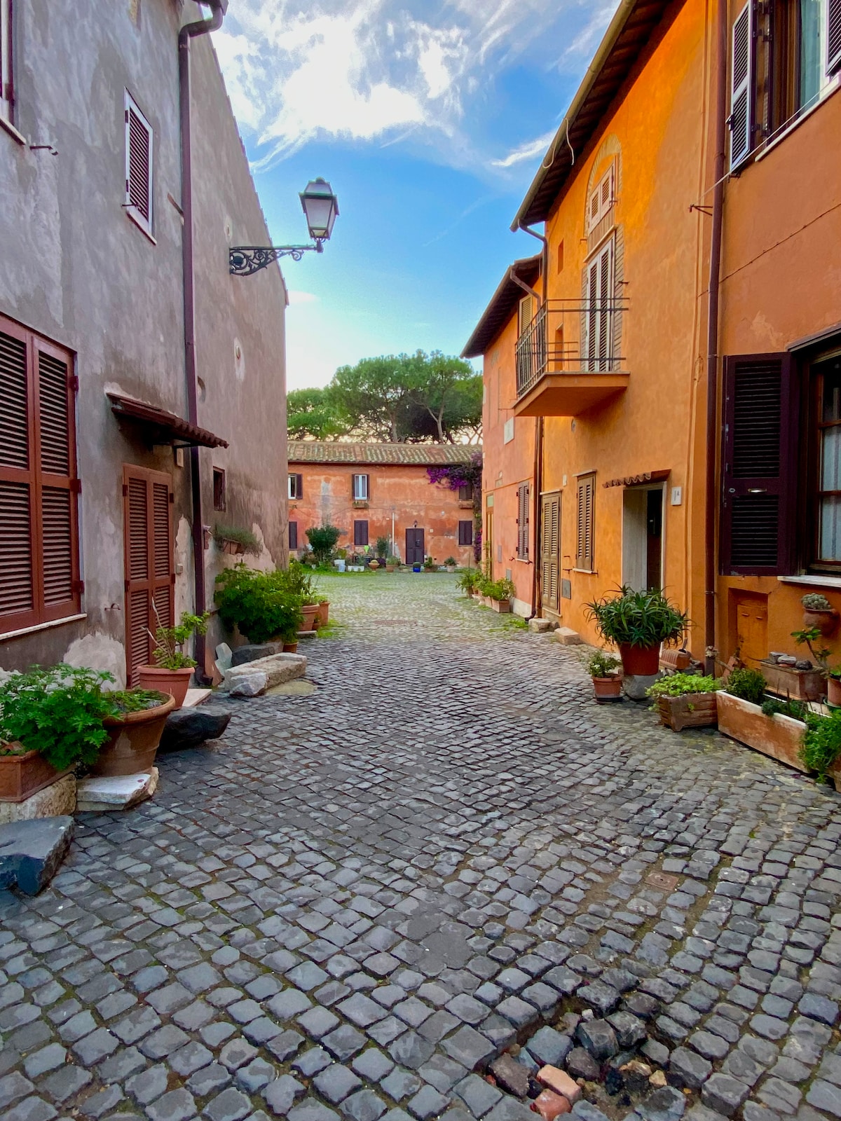 A cobblestone street is flanked by charming buildings in warm, earthy tones. Potted plants adorn the base of windows and doorways, contributing to the inviting atmosphere. A gentle light illuminates the path, while greenery can be seen in the background, suggesting a peaceful outdoor space.