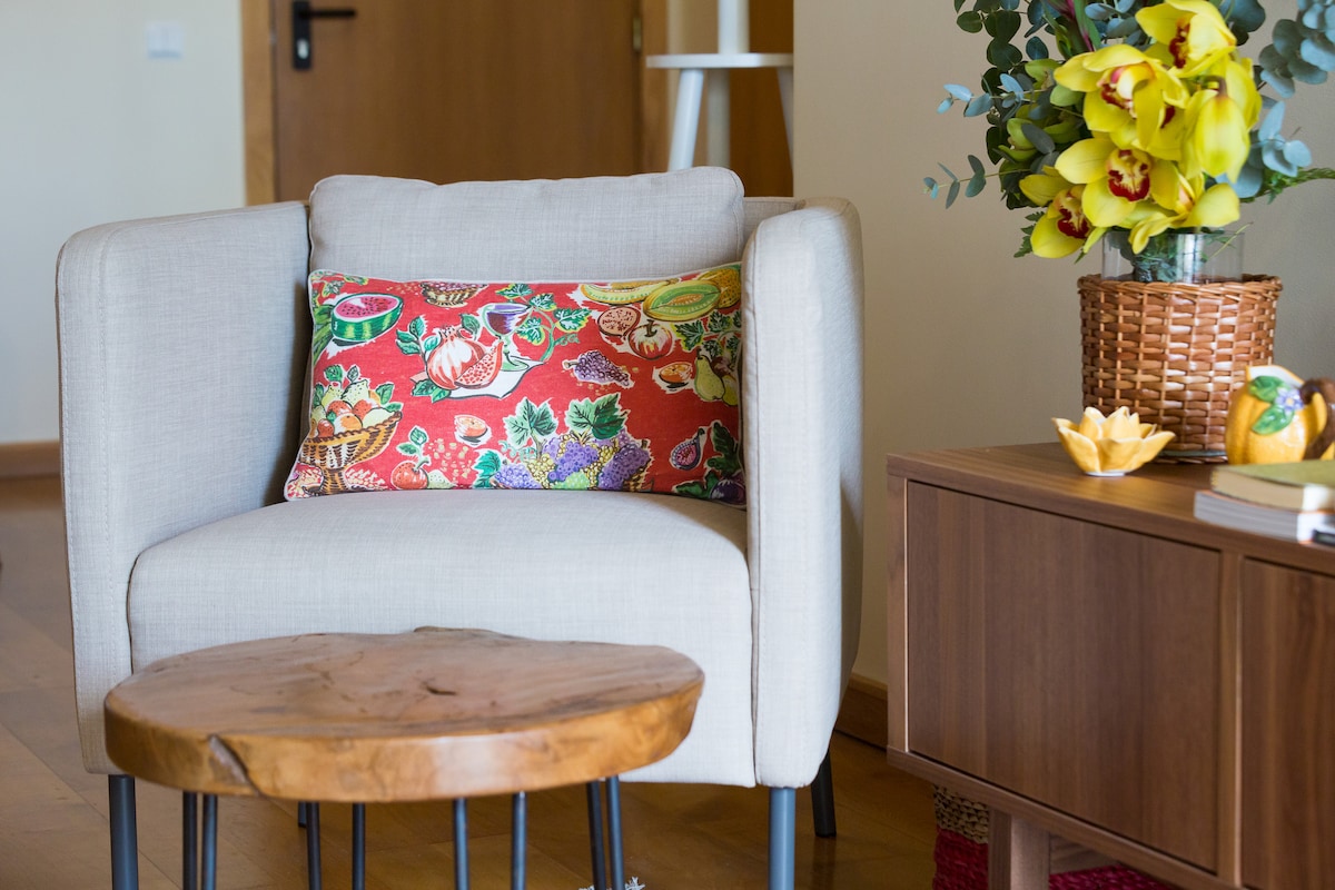 A comfortable armchair featuring a colorful floral cushion is positioned beside a wooden side table. The side table showcases a decorative bowl and a small plant. A cabinet with a warm wood finish is visible, adding to the inviting atmosphere of the living space.