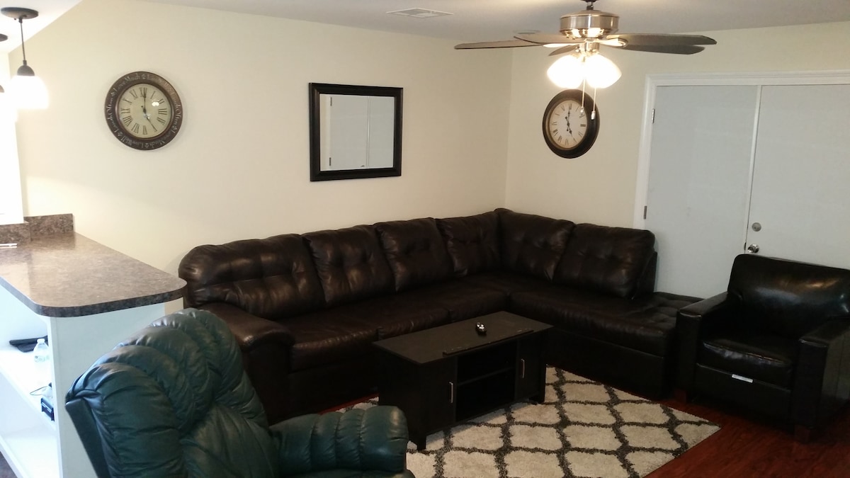 A spacious living room features a large sectional couch alongside a comfortable armchair. A coffee table is placed in the center, with decorative wall clocks adding a touch of character. The area is illuminated by overhead lighting, and wooden flooring enhances the inviting feel.